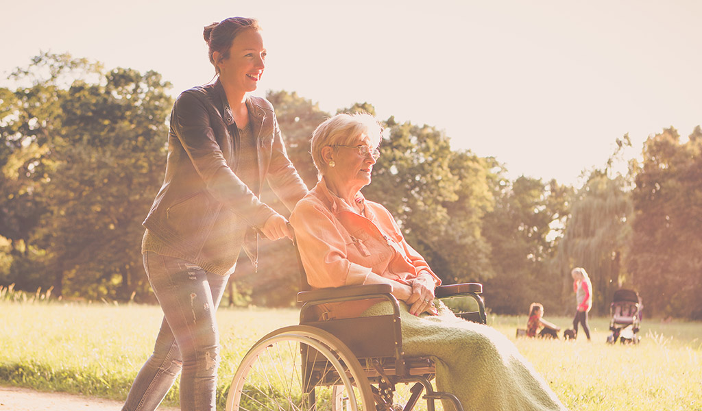 caretaker and older woman in wheelchair walking in a park