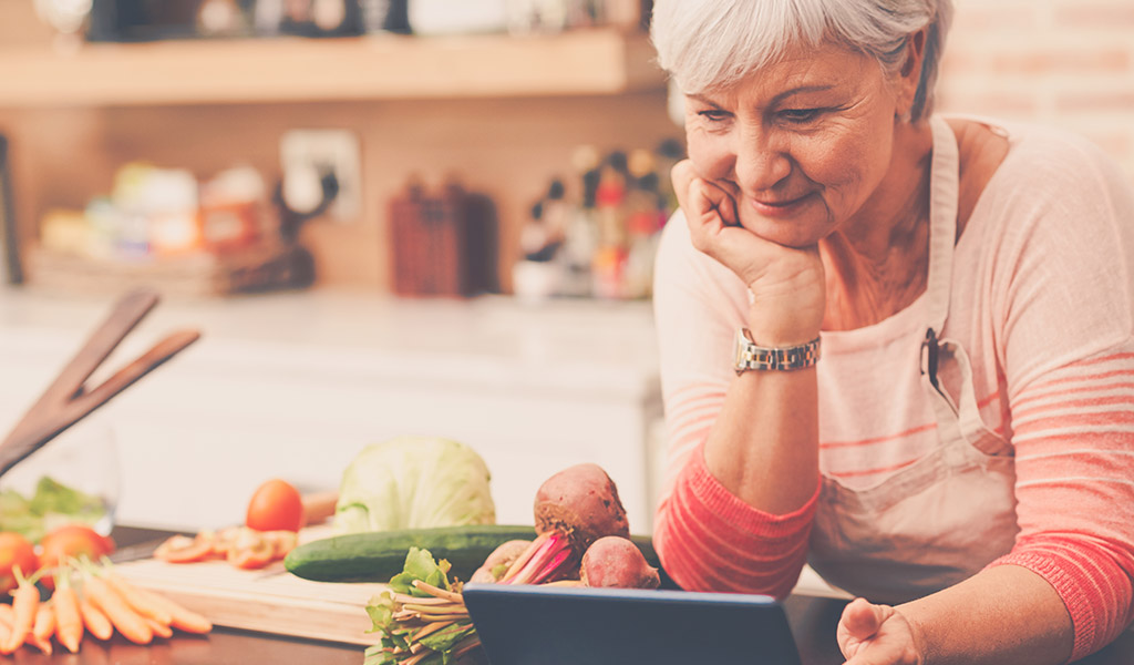 Senior woman looking at tablet for a recipe while standing next to kitchen island covered in vegetables