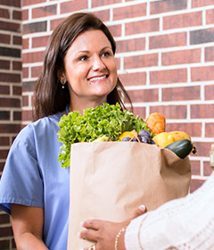 Smiling female caregiver standing at front door holding paper bag filled with groceries