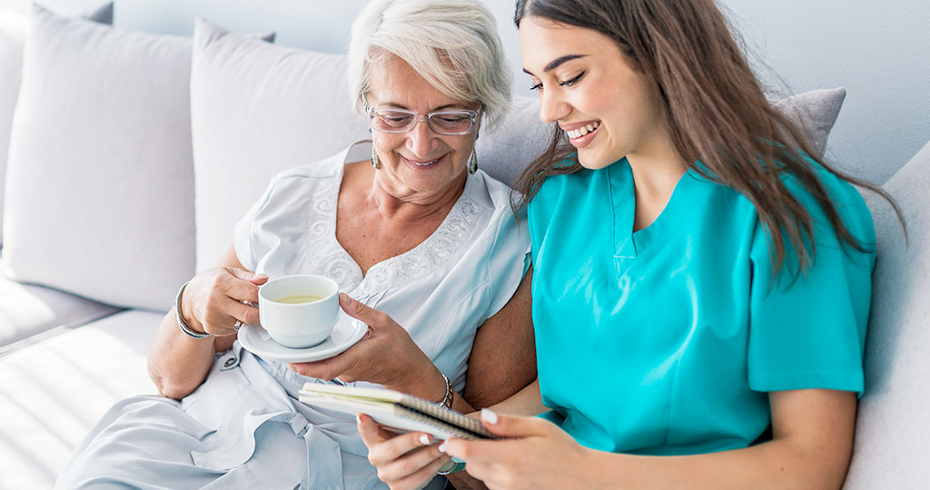 Female caregiver wearing scrubs sitting on couch with senior woman drinking tea and looking at notebook