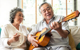 Senior man playing guitar joined by wife singing while sitting on couch