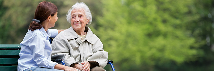 Smiling caregiver sitting on park bench next to happy senior woman in wheelchair in a park