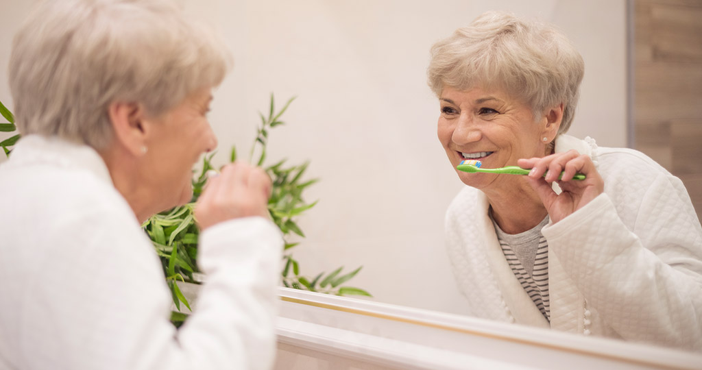 Senior woman standing in front of bathroom mirror while brushing her teeth